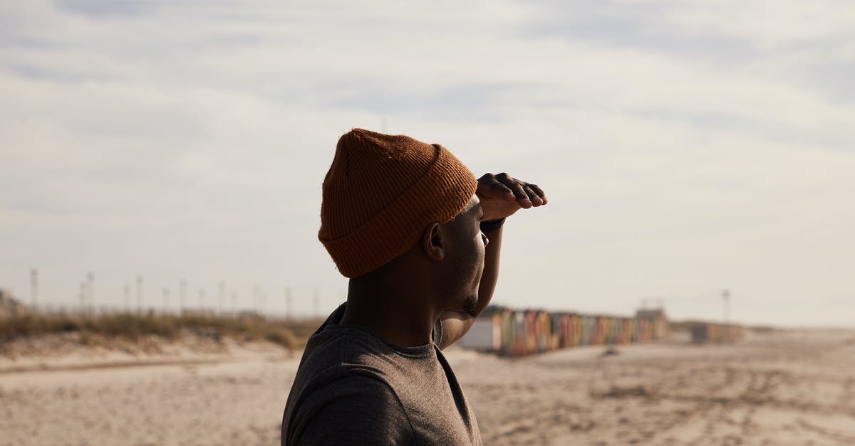 A trip to Patalpani near Indore - Side view of African American male in hat with hand near forehead standing on sandy shore with traces against cloudy sky A trip to Patalpani near Indore - Side view of African American male in hat with hand near forehead standing on sandy shore with traces against cloudy sky