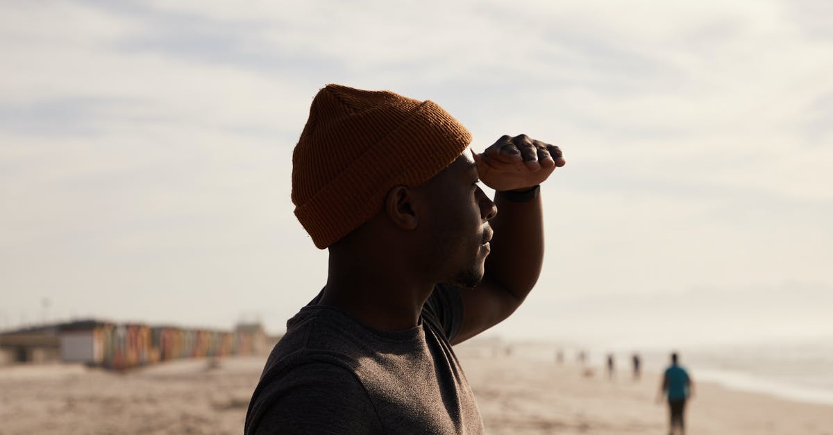 A trip to Patalpani near Indore - Side view of African American male standing with head near forehead on sandy beach near sea with blurred people against cloudy sky A trip to Patalpani near Indore - Side view of African American male standing with head near forehead on sandy beach near sea with blurred people against cloudy sky