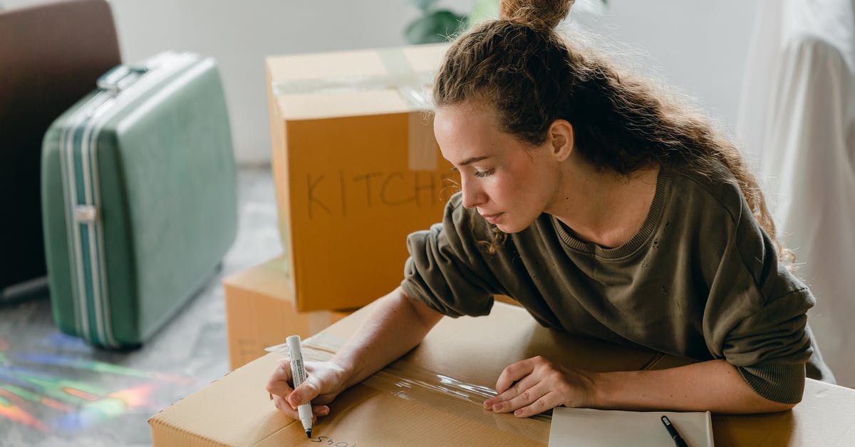 Applying for new B1/B2 visa with new passport, the birthday on my new is different from my old - Top view of thoughtful lady in casual wear writing with marker on cardboard box while sitting near suitcases and pile of boxes in light living room