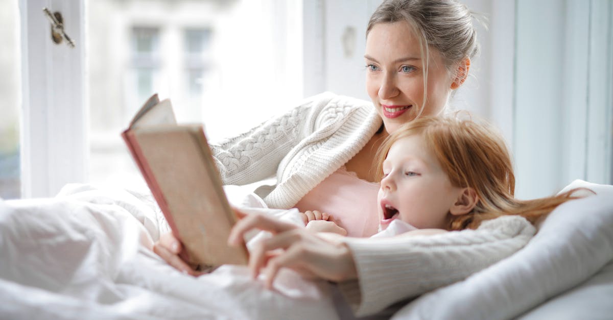 Booking domestic flights in Iran - Cheerful young woman hugging cute little girl and reading book together while lying in soft bed in light bedroom at home in daytime Booking domestic flights in Iran - Cheerful young woman hugging cute little girl and reading book together while lying in soft bed in light bedroom at home in daytime