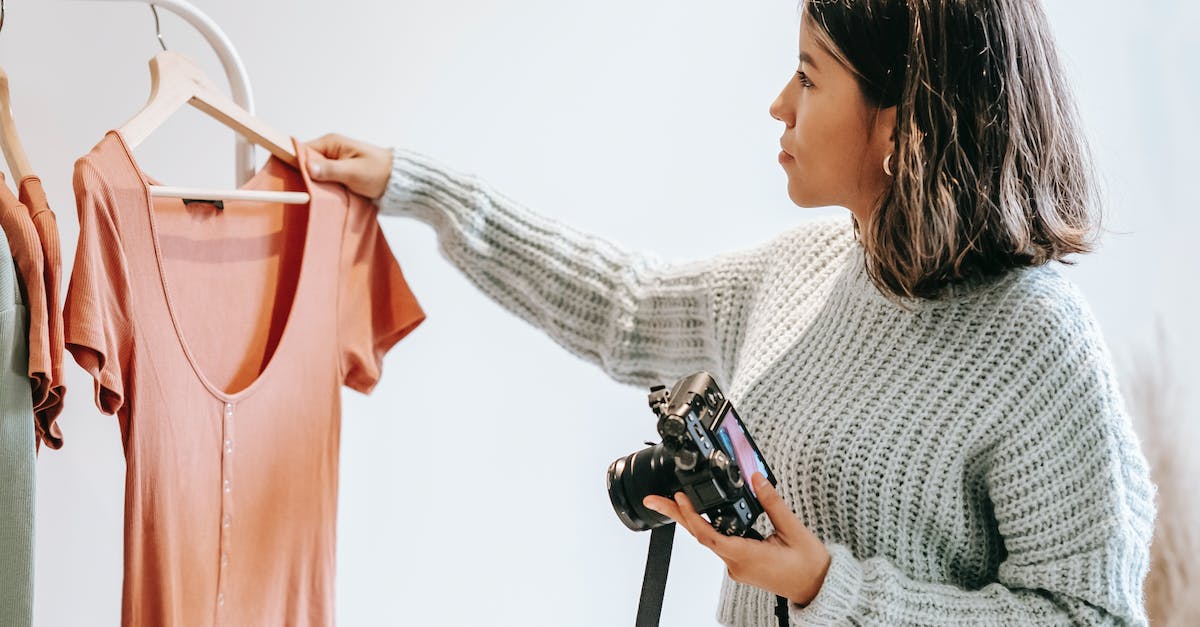 Camera store near Split, Croatia? - Side view of young female in sweater selecting shirt from hanger for photoshoot while holding camera in light studio Camera store near Split, Croatia? - Side view of young female in sweater selecting shirt from hanger for photoshoot while holding camera in light studio