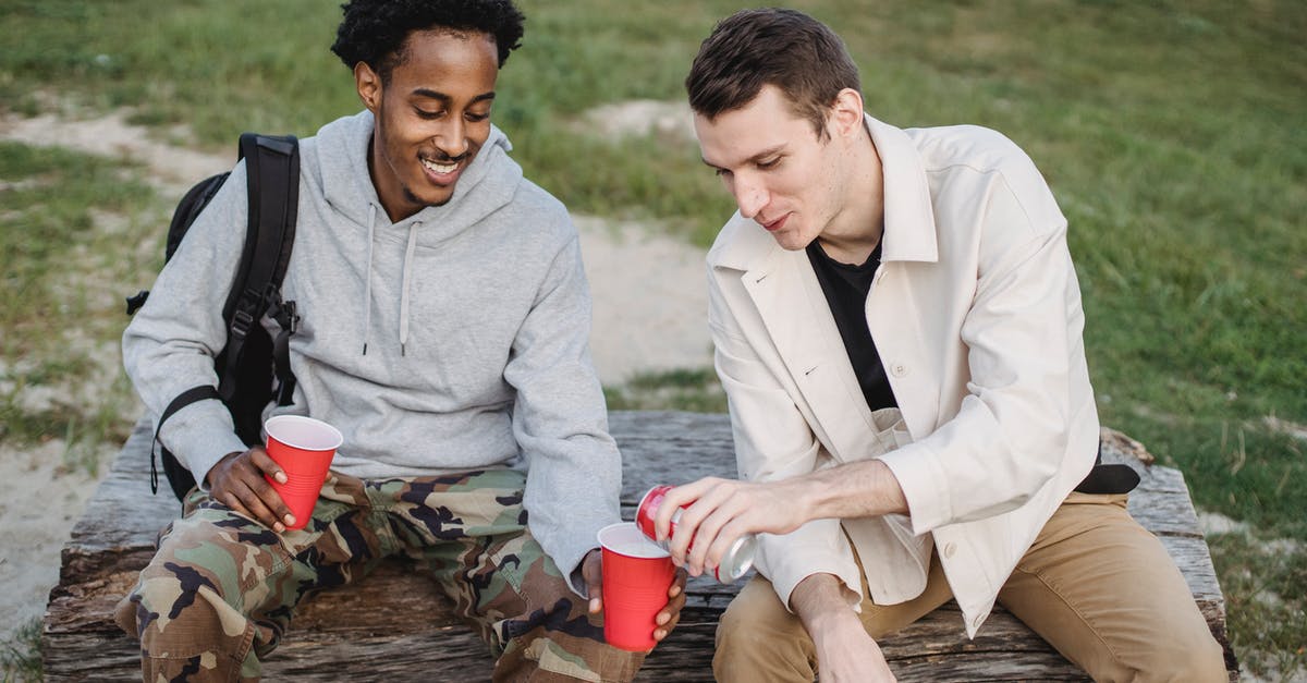 Can I invite a friend to visit me in the UK? - High angle of male pouring carbonated drink from tin can to smiling black friend sitting with red cup Can I invite a friend to visit me in the UK? - High angle of male pouring carbonated drink from tin can to smiling black friend sitting with red cup