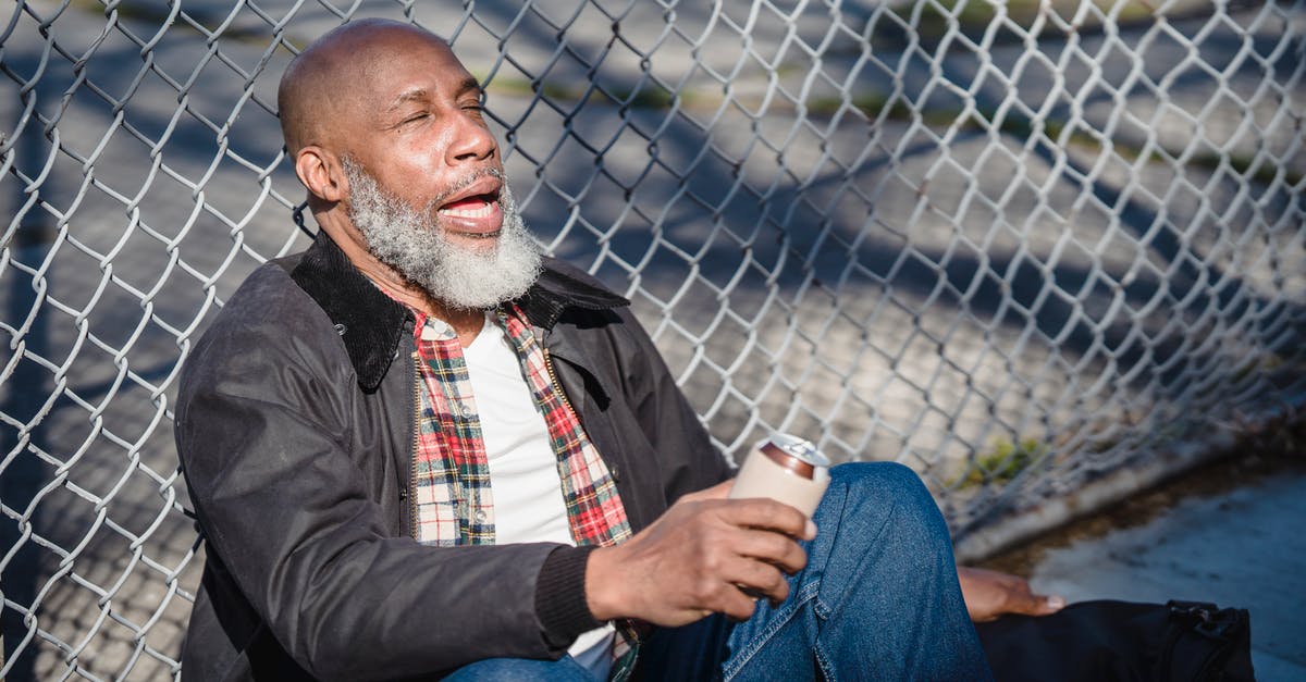 Can I sleep overnight at Stansted Airport? - African American senior man with closed eyes and opened mouth sitting near metal fence and holding beer can Can I sleep overnight at Stansted Airport? - African American senior man with closed eyes and opened mouth sitting near metal fence and holding beer can
