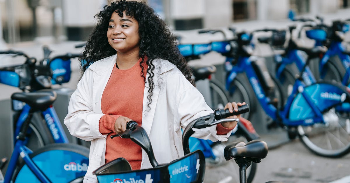 Can you rent a bicycle in a city and return it in another one in France? - Cheerful African American female bicyclist in casual clothes renting bicycle in bicycle sharing station on city street and looking away contentedly Can you rent a bicycle in a city and return it in another one in France? - Cheerful African American female bicyclist in casual clothes renting bicycle in bicycle sharing station on city street and looking away contentedly
