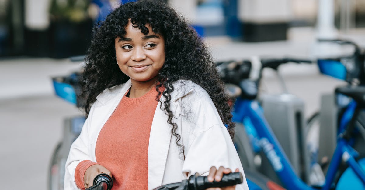 Can you rent a bicycle in a city and return it in another one in France? - Smiling African American female cyclist wearing casual outfit standing with bicycle near bicycle sharing station on modern city street Can you rent a bicycle in a city and return it in another one in France? - Smiling African American female cyclist wearing casual outfit standing with bicycle near bicycle sharing station on modern city street
