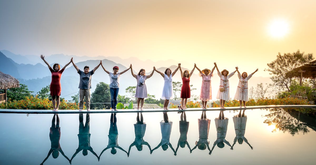 Can you share frequent flyer status with people in your travel group? - Happy full body ethnic people in summer clothes standing on poolside and holding hands with raised arms near calm water on sunset with amazing mountains on background