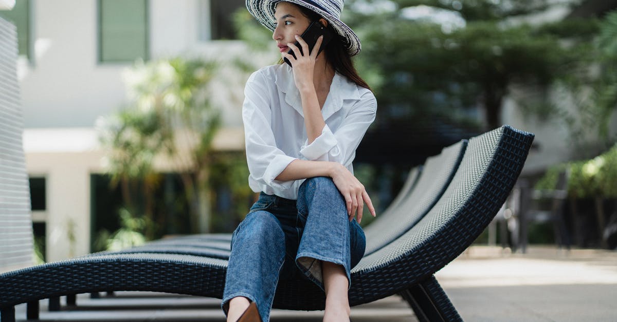 Cancellation fee for a hotel in Austria booked via phone call - Full body of young stylish ethnic female in high heeled shoes and hat sitting on sunbed and having phone conversation in hotel garden Cancellation fee for a hotel in Austria booked via phone call - Full body of young stylish ethnic female in high heeled shoes and hat sitting on sunbed and having phone conversation in hotel garden