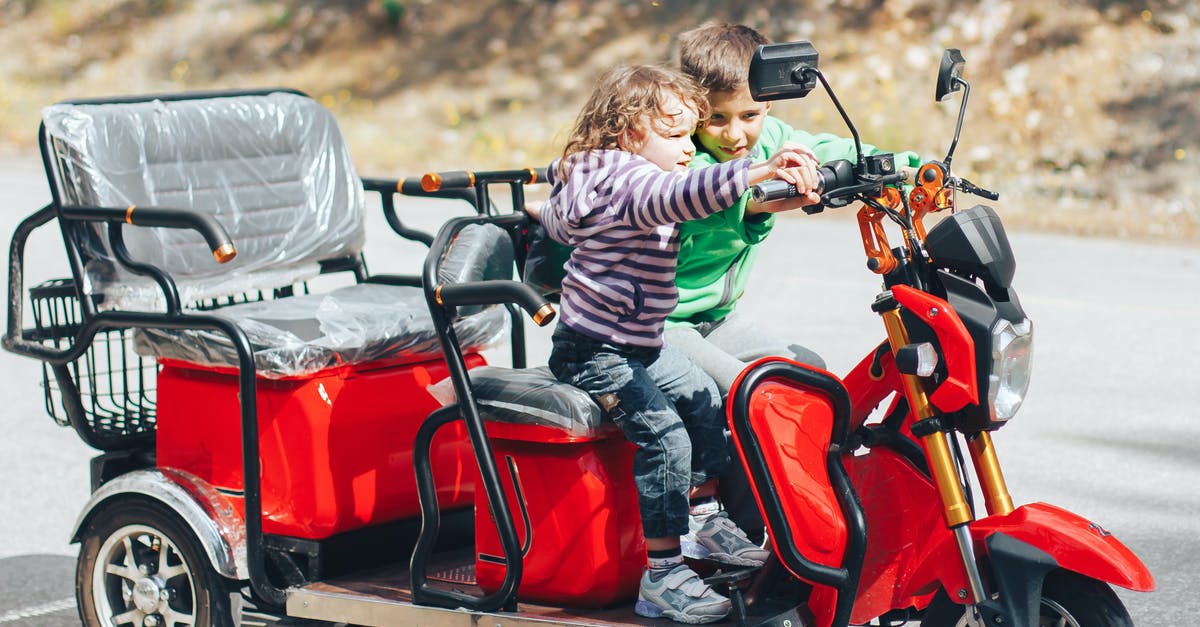 Cheap and convenient way to travel with kids around US - Adorable child helping little brother to ride modern red trike motorcycle on asphalt road on sunny day Cheap and convenient way to travel with kids around US - Adorable child helping little brother to ride modern red trike motorcycle on asphalt road on sunny day