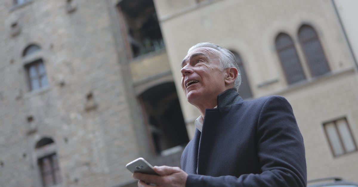 Check in to 2 hotels at same location - From below of mature gentleman in classy wear standing near building in city and checking message on cellphone while looking up Check in to 2 hotels at same location - From below of mature gentleman in classy wear standing near building in city and checking message on cellphone while looking up