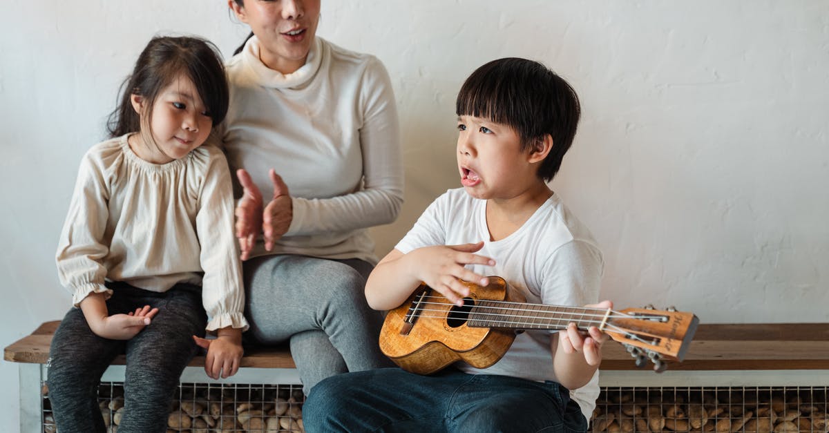 Children of U.K. Passport holder on accompaniment visas - Ethnic boy making grimace while playing ukulele for mother Children of U.K. Passport holder on accompaniment visas - Ethnic boy making grimace while playing ukulele for mother