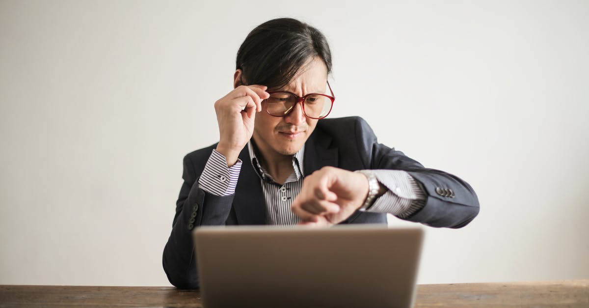 Connecting time ay JFK - Young frowning man in suit and glasses looking at wristwatch while waiting for appointment sitting at desk with laptop Connecting time ay JFK - Young frowning man in suit and glasses looking at wristwatch while waiting for appointment sitting at desk with laptop