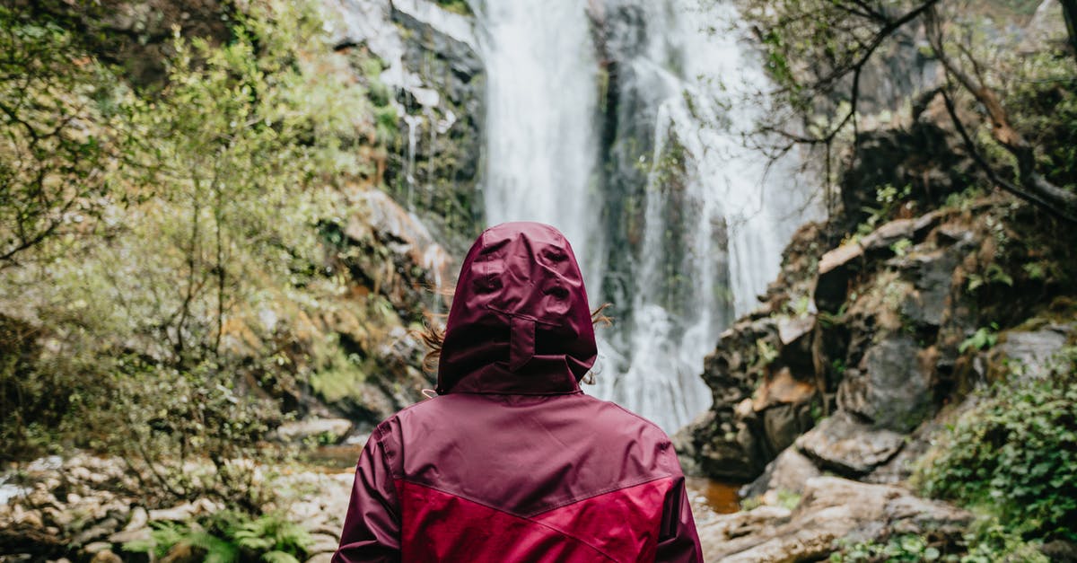 Do the Canadian Rockies lack red foliage in autumn? - Back view of anonymous tourist in red outerwear with hood standing near flowing stream of water falling from rocky cliff in daylight Do the Canadian Rockies lack red foliage in autumn? - Back view of anonymous tourist in red outerwear with hood standing near flowing stream of water falling from rocky cliff in daylight