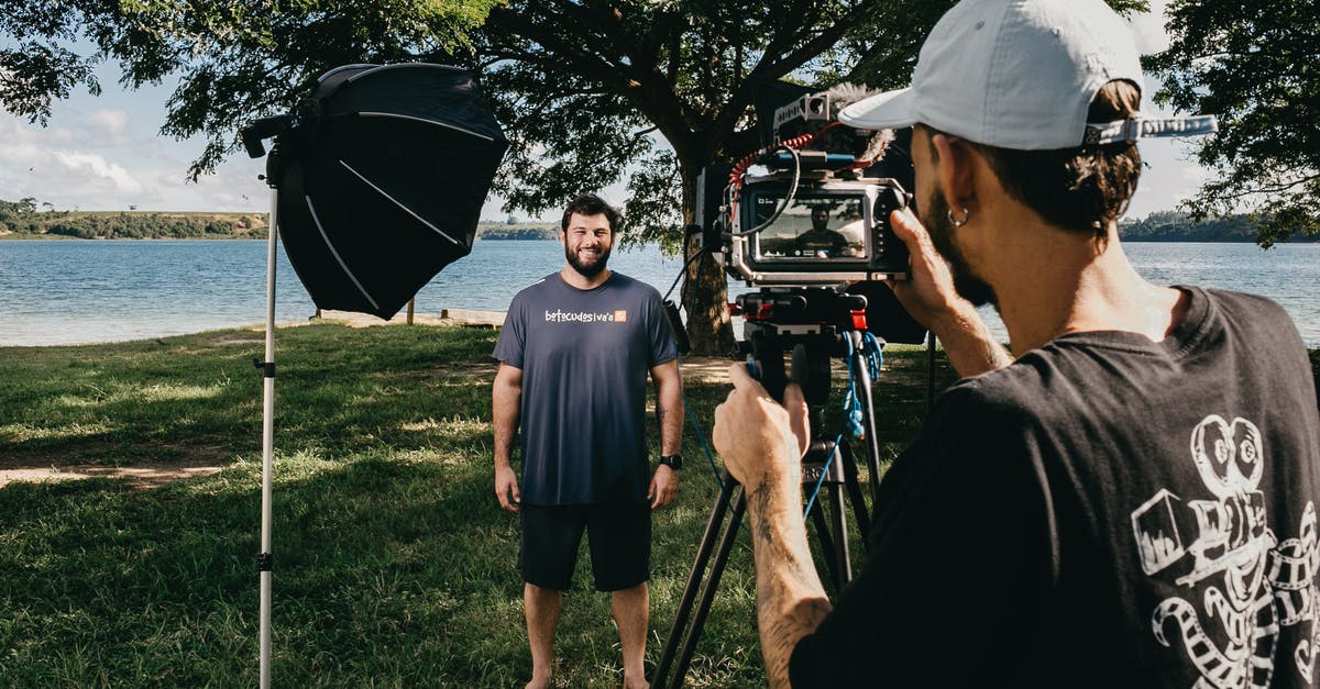 Do UK Immigration Officers record video when interviewing arrivers? - Filmmaker with video camera on tripod shooting video of smiling man standing on glade on coast of pond
