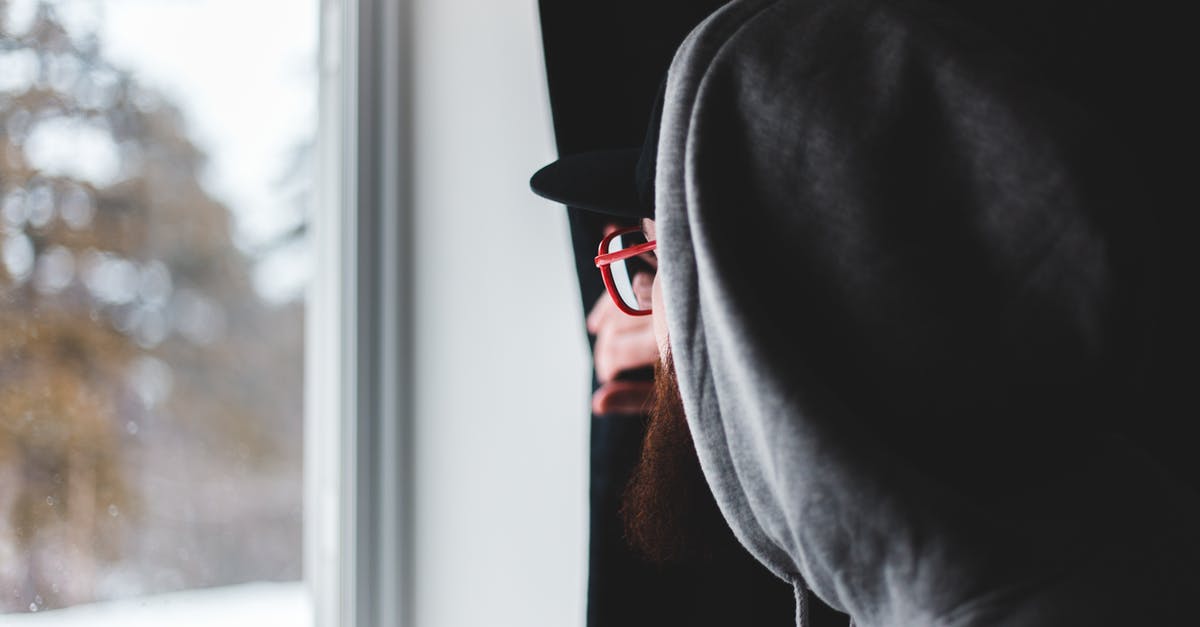 Domestic Or International Check In - Side view of bearded man in glasses and hoodie pulling curtain and looking out window on winter day at home Domestic Or International Check In - Side view of bearded man in glasses and hoodie pulling curtain and looking out window on winter day at home