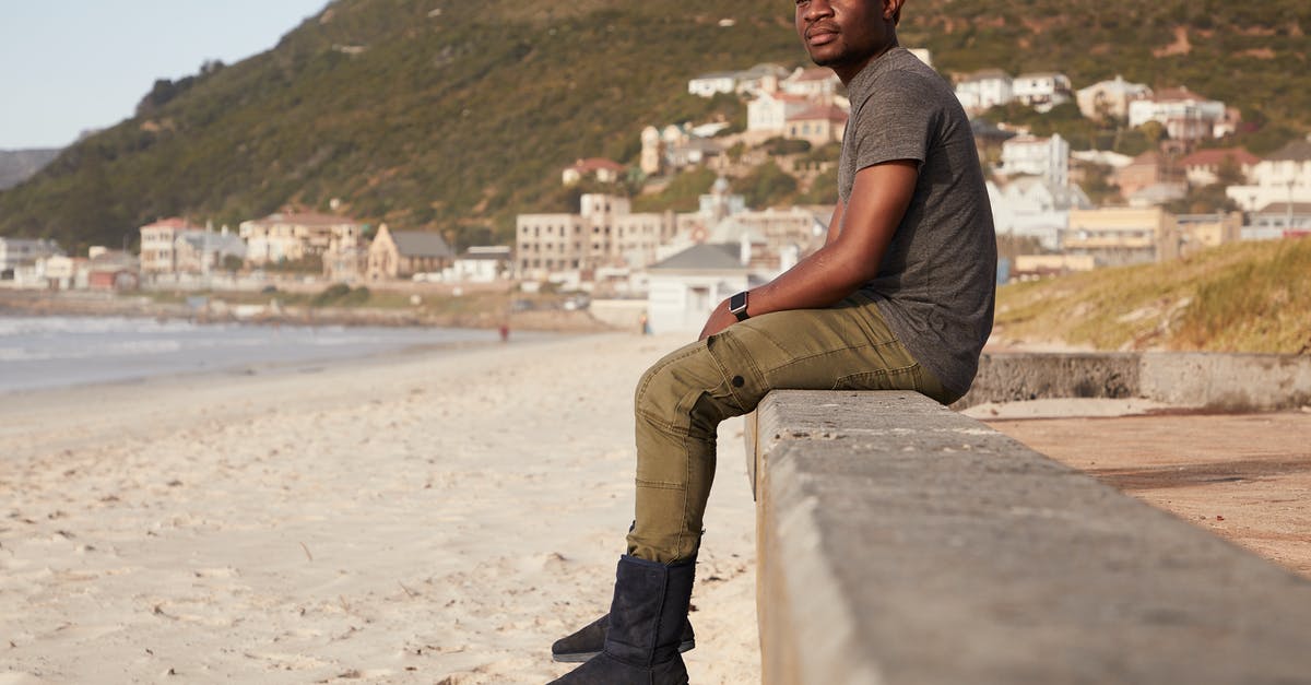 Dual Citizen Border Hopping to Extend Tourist Visa - Full body of thoughtful African American male in hat sitting on stone border on sandy coast near sea with coastal town in distance