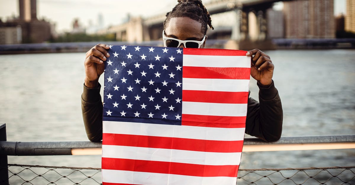 Dual citizen (Tunisia/Canada) travelling to USA - Ethnic male in casual clothes and sunglasses standing on embankment of city river while leaning on fence showing national flag of United States of America Dual citizen (Tunisia/Canada) travelling to USA - Ethnic male in casual clothes and sunglasses standing on embankment of city river while leaning on fence showing national flag of United States of America