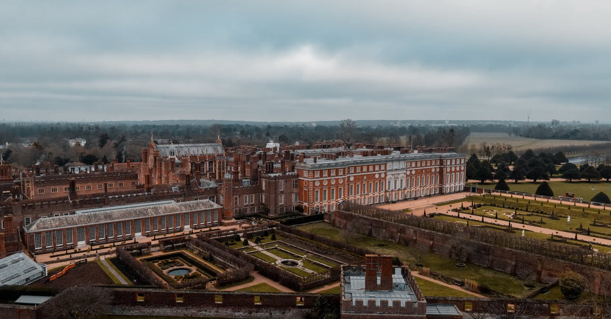 Extending 7 day London Oyster card for extra zones - Drone view of Hampton Court Palace located in England in London with green grassy lawns with plants near fence and trees near pathways with people under cloudy gray sky Extending 7 day London Oyster card for extra zones - Drone view of Hampton Court Palace located in England in London with green grassy lawns with plants near fence and trees near pathways with people under cloudy gray sky