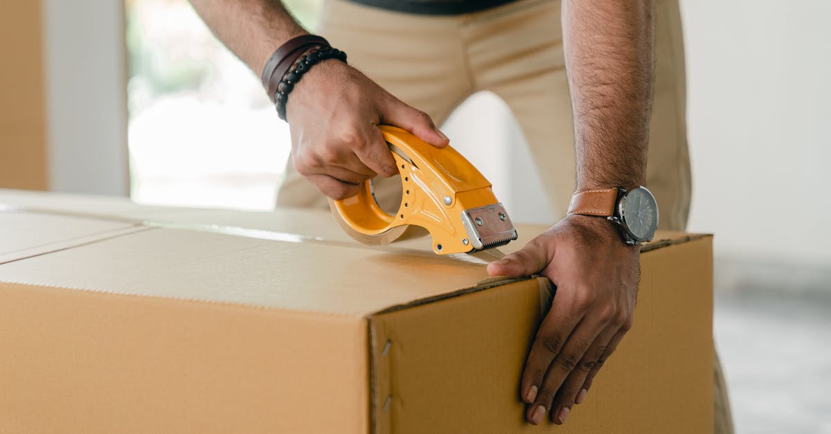 Foreign storage of a carry-on - Crop faceless young male with wristwatch using adhesive tape while preparing cardboard box for transportation Foreign storage of a carry-on - Crop faceless young male with wristwatch using adhesive tape while preparing cardboard box for transportation