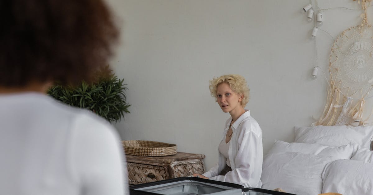 Free extra baggage with trans-Atlantic flights with KLM? - Content lady with blond hair preparing to leave while sitting on bed near white wall in bedroom with crop female friend with dark hair Free extra baggage with trans-Atlantic flights with KLM? - Content lady with blond hair preparing to leave while sitting on bed near white wall in bedroom with crop female friend with dark hair