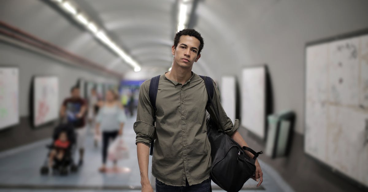 Getting from San Francisco (Union Square) to Oakland Airport by public transport - Calm young African American male in casual clothes with big black bag and backpack looking at camera while walking along corridor of underground station against blurred passengers