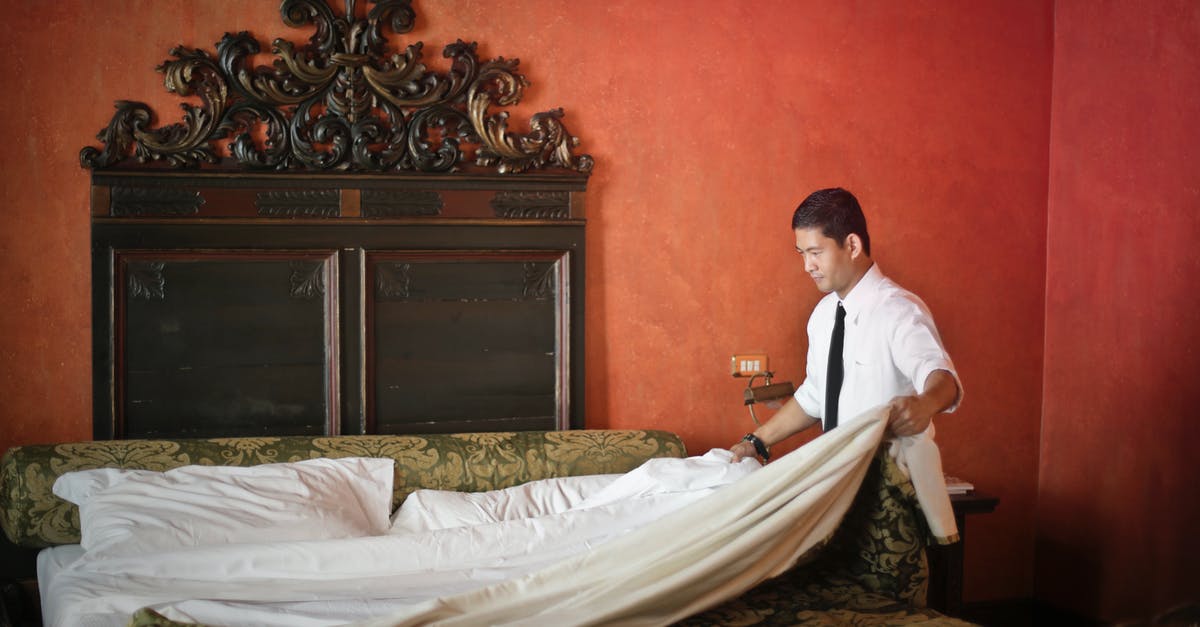 Hotels in Barcelona for People Aged 17? - Side view of young ethnic male making bed while cleaning stylish hotel room with vintage furniture and red walls Hotels in Barcelona for People Aged 17? - Side view of young ethnic male making bed while cleaning stylish hotel room with vintage furniture and red walls
