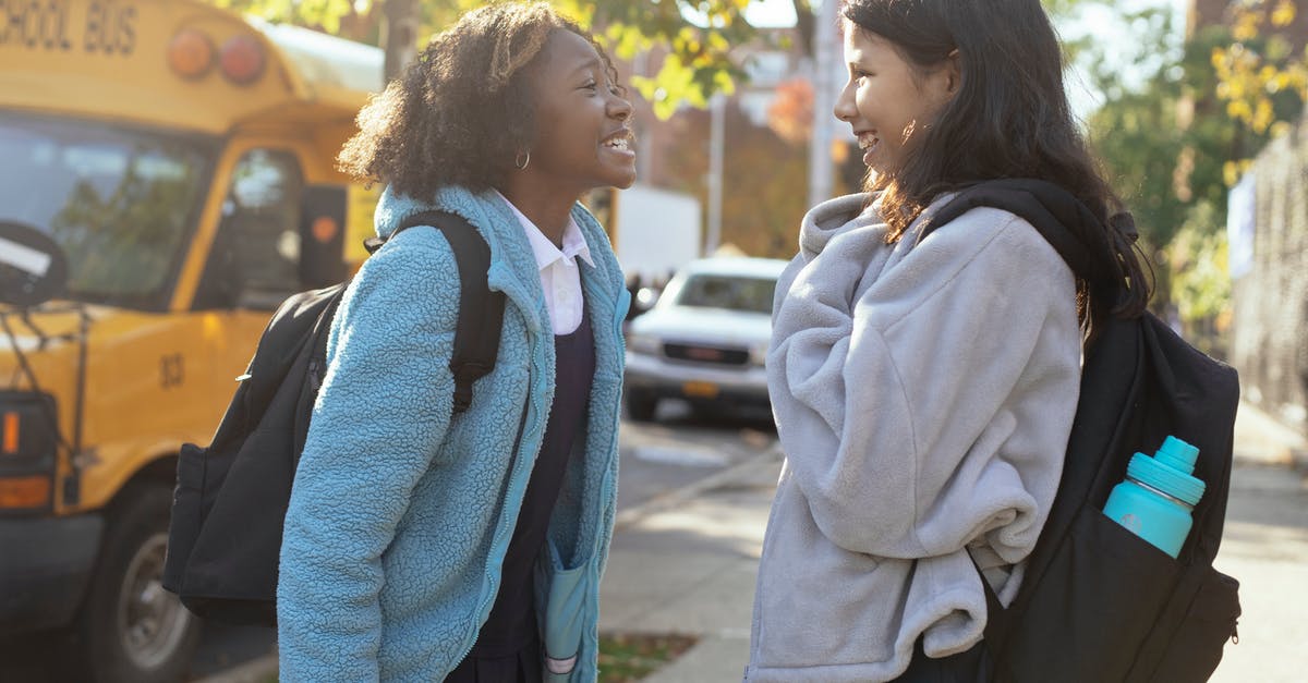How do US intercity bus companies relate to each other? - Side view happy multiracial schoolgirls in warm jackets with backpacks standing on street near school bus and talking while looking at each other with smile How do US intercity bus companies relate to each other? - Side view happy multiracial schoolgirls in warm jackets with backpacks standing on street near school bus and talking while looking at each other with smile