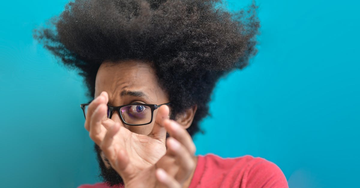How does airport security search through one's luggage? - Scared young ethnic man looking through hands on blue background in studio How does airport security search through one's luggage? - Scared young ethnic man looking through hands on blue background in studio