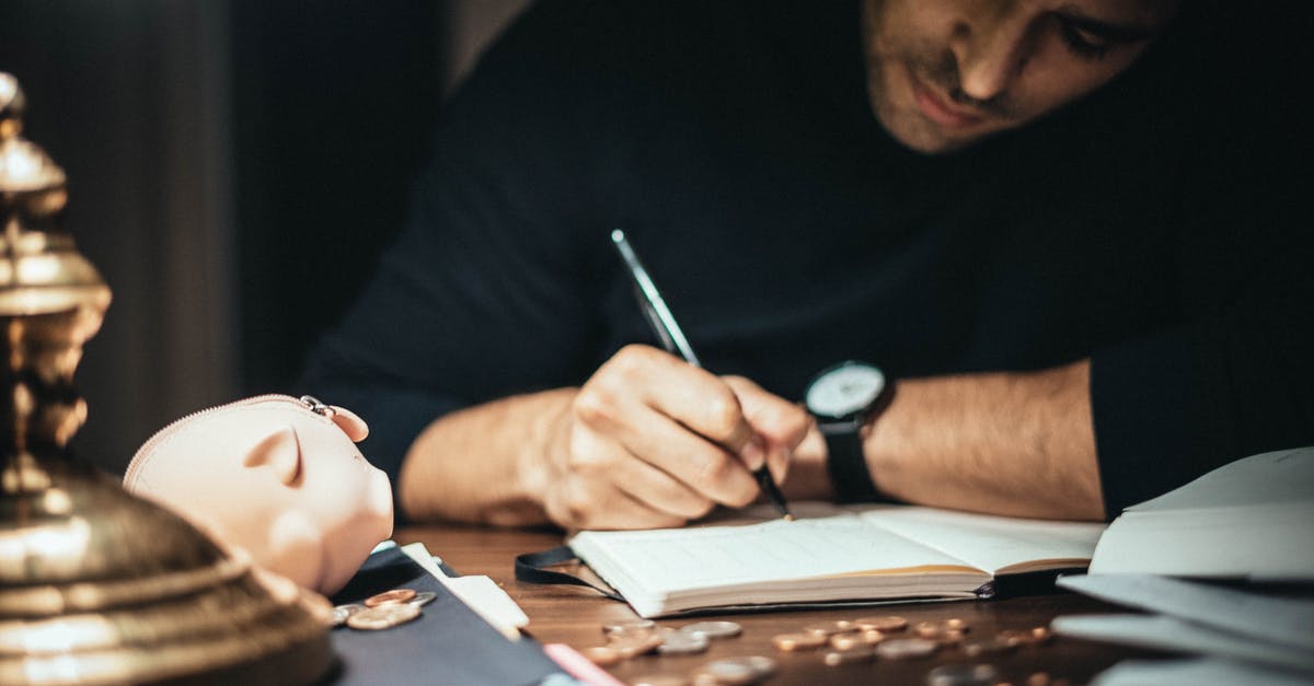 How to take money to Iran? - Crop elegant man taking notes in journal while working at desk with coins and piggybank in lamplight How to take money to Iran? - Crop elegant man taking notes in journal while working at desk with coins and piggybank in lamplight