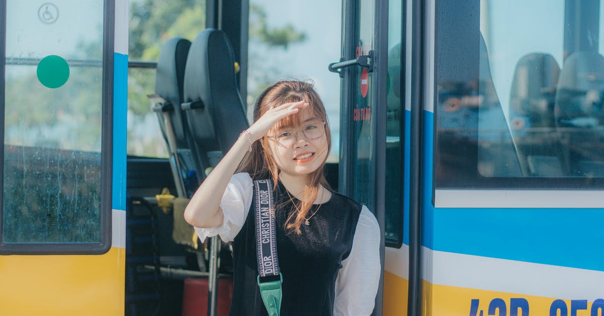 I want to take a bus from Dusseldorf Airport to NH Hotel City Centre - please tell me what would be the bus number - Young cheerful slim Asian passenger with shoulder bag in eyewear touching head while looking at camera after getting off colorful bus in city I want to take a bus from Dusseldorf Airport to NH Hotel City Centre - please tell me what would be the bus number - Young cheerful slim Asian passenger with shoulder bag in eyewear touching head while looking at camera after getting off colorful bus in city
