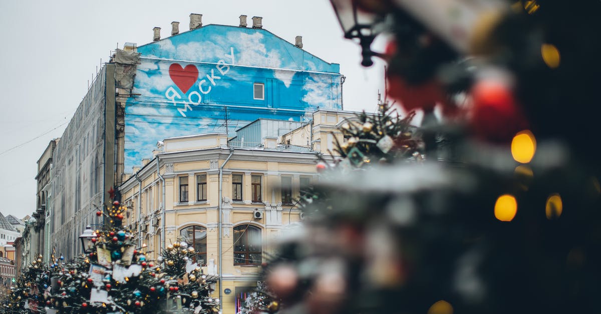 Identify the building in this picture - Facade of old building with creative graffiti with inscription located on city square with Christmas trees decorated with ornaments and colorful baubles Identify the building in this picture - Facade of old building with creative graffiti with inscription located on city square with Christmas trees decorated with ornaments and colorful baubles