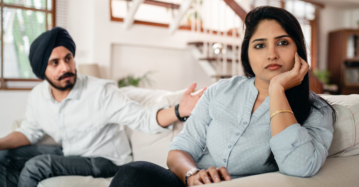 Indian eBusiness visa issues - Young bearded Sikh man in casual wear and turban arguing with sad wife sitting on sofa Indian eBusiness visa issues - Young bearded Sikh man in casual wear and turban arguing with sad wife sitting on sofa