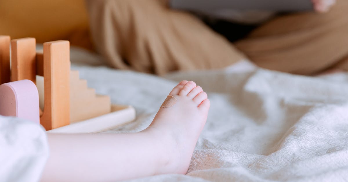 Infant visa online questionaire - Crop faceless toddler sitting on bed with colorful wooden building blocks while mother sitting with legs crossed doing remote work with laptop on knees on background Infant visa online questionaire - Crop faceless toddler sitting on bed with colorful wooden building blocks while mother sitting with legs crossed doing remote work with laptop on knees on background
