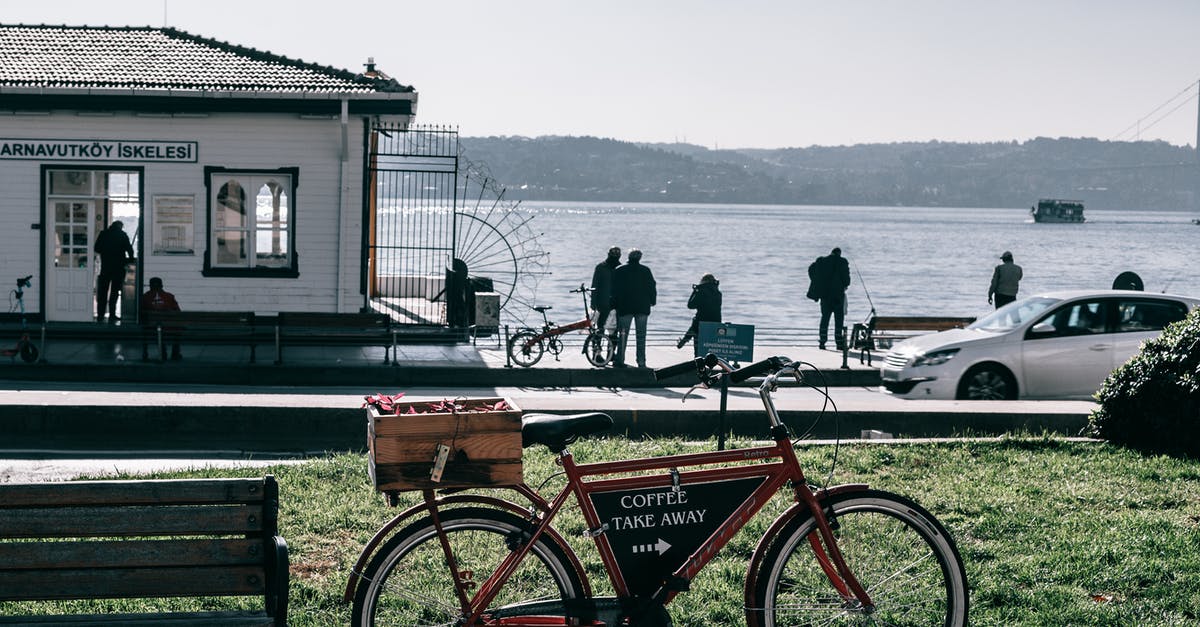 Information about public transport in Greece - Bicycle with pointer on black board parked on crowded paved embankment with car and building against calm sea and mountain on background Information about public transport in Greece - Bicycle with pointer on black board parked on crowded paved embankment with car and building against calm sea and mountain on background