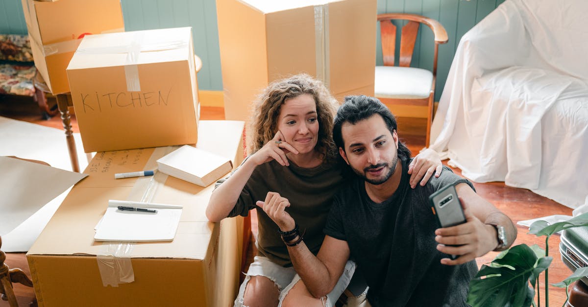 Is it mandatory to show my credit card upon my arrival at hotel booked using Booking website? - Top view of trendy woman embracing ethnic boyfriend in casual wear while taking selfie on cellphone and sitting surrounded by heap of cardboard boxes near sofa covered with bed sheet