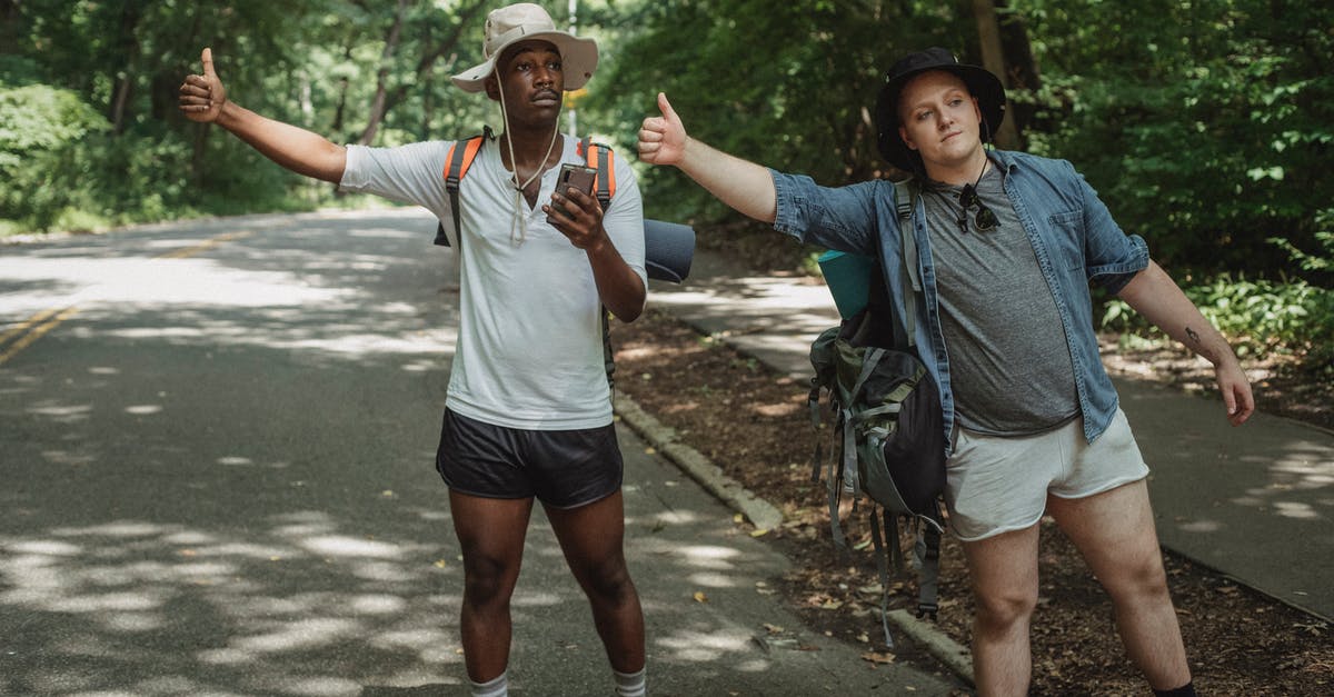 Is it possible to hitchhike to Galapagos? - Young multiethnic tourists with backpacks standing with smartphone while showing like gesture on roadway near forest and looking away Is it possible to hitchhike to Galapagos? - Young multiethnic tourists with backpacks standing with smartphone while showing like gesture on roadway near forest and looking away