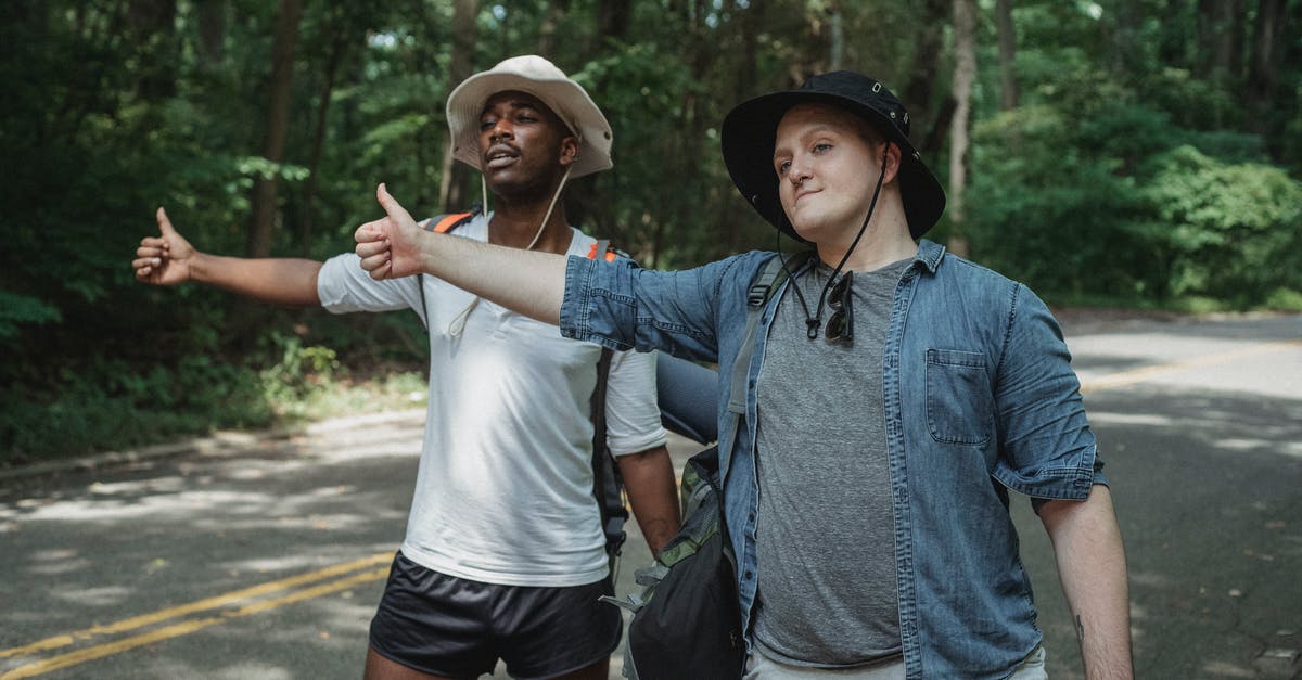 Is it possible to hitchhike to Galapagos? - Content multiethnic male hitchhikers in casual outfits and hats with backpacks standing on roadside with thumbs up in lush forest Is it possible to hitchhike to Galapagos? - Content multiethnic male hitchhikers in casual outfits and hats with backpacks standing on roadside with thumbs up in lush forest
