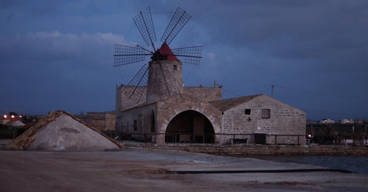 Is it possible to visit a water pumping windmill and a industrial windmill at the same location - Aged stone building with windmill placed on concrete surface near river under cloudy sky in evening Is it possible to visit a water pumping windmill and a industrial windmill at the same location - Aged stone building with windmill placed on concrete surface near river under cloudy sky in evening
