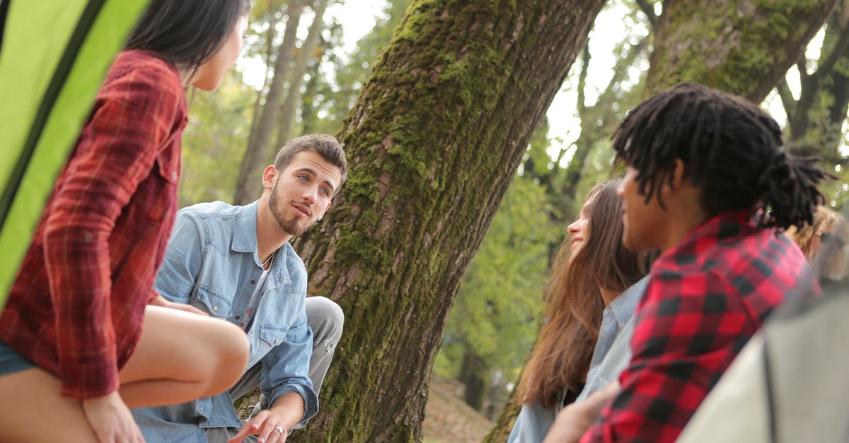 Is it safe to make groups/families sit apart from each other? - Group of young people in casual clothes sitting on plaid around near trees with moss while talking and looking at each other Is it safe to make groups/families sit apart from each other? - Group of young people in casual clothes sitting on plaid around near trees with moss while talking and looking at each other