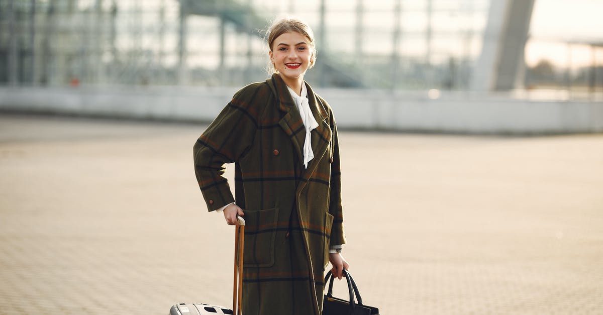 Lay over at El Prat Barcelona, luggage and city tour - Positive smiling young female traveler in casual warm coat with suitcase and ladies bag standing near airport terminal and looking at camera Lay over at El Prat Barcelona, luggage and city tour - Positive smiling young female traveler in casual warm coat with suitcase and ladies bag standing near airport terminal and looking at camera