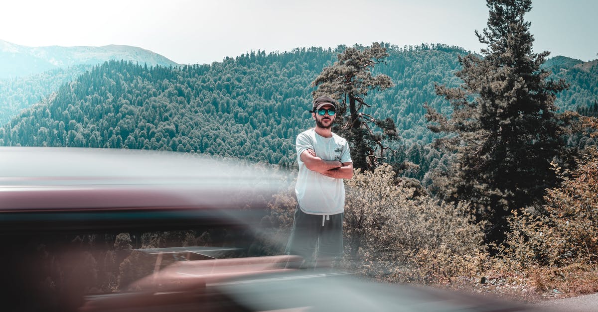 Long distance (16 hours) car trip with rental car and driver - Long exposure of male in casual clothing standing between mountains covered with woodland and car riding across road Long distance (16 hours) car trip with rental car and driver - Long exposure of male in casual clothing standing between mountains covered with woodland and car riding across road