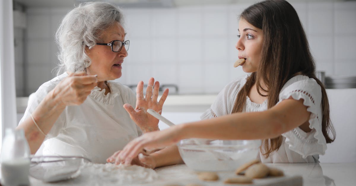 Looking for hurricane advice and sources - Calm senior woman and teenage girl in casual clothes looking at each other and talking while eating cookies and cooking pastry in contemporary kitchen at home