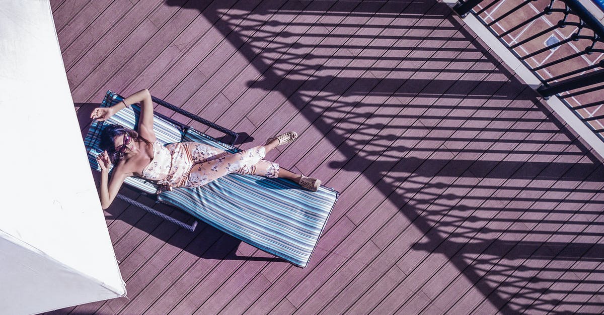 Melbourne: Traveling from Southern Cross station to hotel at 11 PM - Top view of anonymous female traveler in sunglasses lying with crossed legs on sun bed on wooden floor near fence of hotel balcony in sunlight during trip Melbourne: Traveling from Southern Cross station to hotel at 11 PM - Top view of anonymous female traveler in sunglasses lying with crossed legs on sun bed on wooden floor near fence of hotel balcony in sunlight during trip