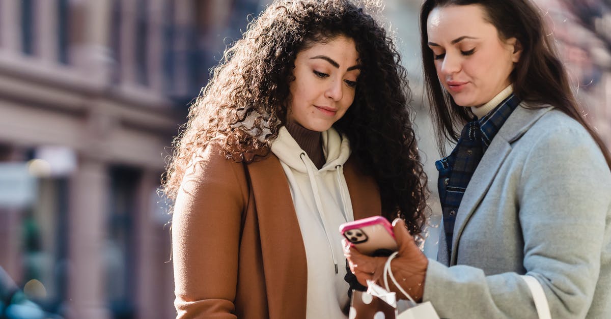 Mobile internet (3G) in UK for some days - Young girlfriends in warm coats and gloves standing in street with gift packages while looking at information on cellphone near buildings in town in sunny day Mobile internet (3G) in UK for some days - Young girlfriends in warm coats and gloves standing in street with gift packages while looking at information on cellphone near buildings in town in sunny day