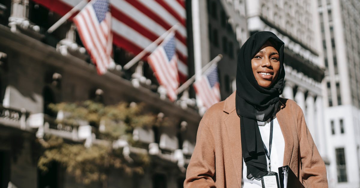 Nearest Iranian embassy for Canadians - From below of cheerful African American female ambassador with folder wearing hijab and id card looking away while standing near building with American flags on blurred background Nearest Iranian embassy for Canadians - From below of cheerful African American female ambassador with folder wearing hijab and id card looking away while standing near building with American flags on blurred background