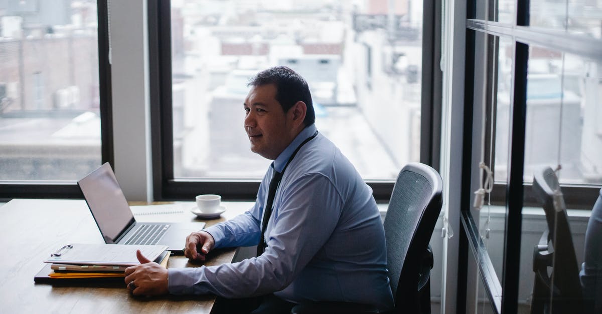 NOC from employer for Schengen visa - High angle side view of smiling ethnic businessman sitting in armchair at table with netbook and documents NOC from employer for Schengen visa - High angle side view of smiling ethnic businessman sitting in armchair at table with netbook and documents