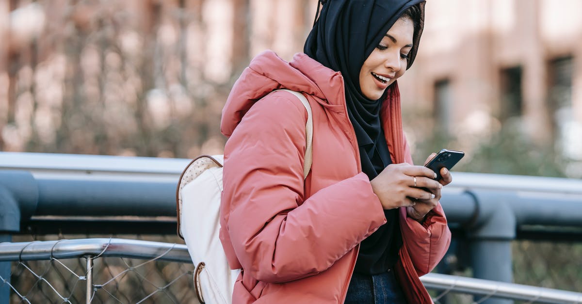 Online check-in not permitted due to my nationality? - Muslim ethnic female in hijab texting message on mobile phone near iron construction on blurred background of building