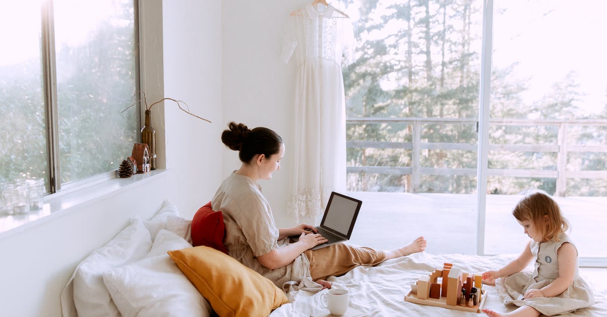 Online resource for duty-free - Side view of cute toddler girl sitting on bed barefoot and playing with colorful wooden blocks while mother using laptop in bed enjoying morning coffee Online resource for duty-free - Side view of cute toddler girl sitting on bed barefoot and playing with colorful wooden blocks while mother using laptop in bed enjoying morning coffee