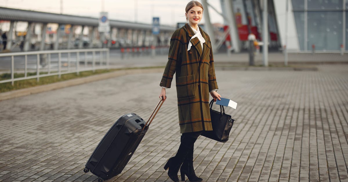 Passport queue length in UK in relation to arrival method - Full body young female passenger in trendy plaid coat with suitcase strolling on pavement near modern building of ariport terminal taking bag and documents Passport queue length in UK in relation to arrival method - Full body young female passenger in trendy plaid coat with suitcase strolling on pavement near modern building of ariport terminal taking bag and documents