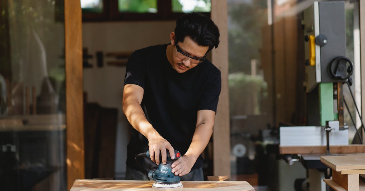 Polish Airports' fingerprint Process - Concentrated male master in protective glasses smoothing wooden plank with sander while creating wooden detail in workroom Polish Airports' fingerprint Process - Concentrated male master in protective glasses smoothing wooden plank with sander while creating wooden detail in workroom