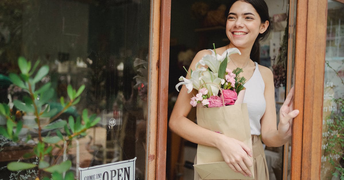 Purchase extra baggage now or wait till checkin time? - Toothy smiling female standing near flower shop and holding paper package with bouquet Purchase extra baggage now or wait till checkin time? - Toothy smiling female standing near flower shop and holding paper package with bouquet